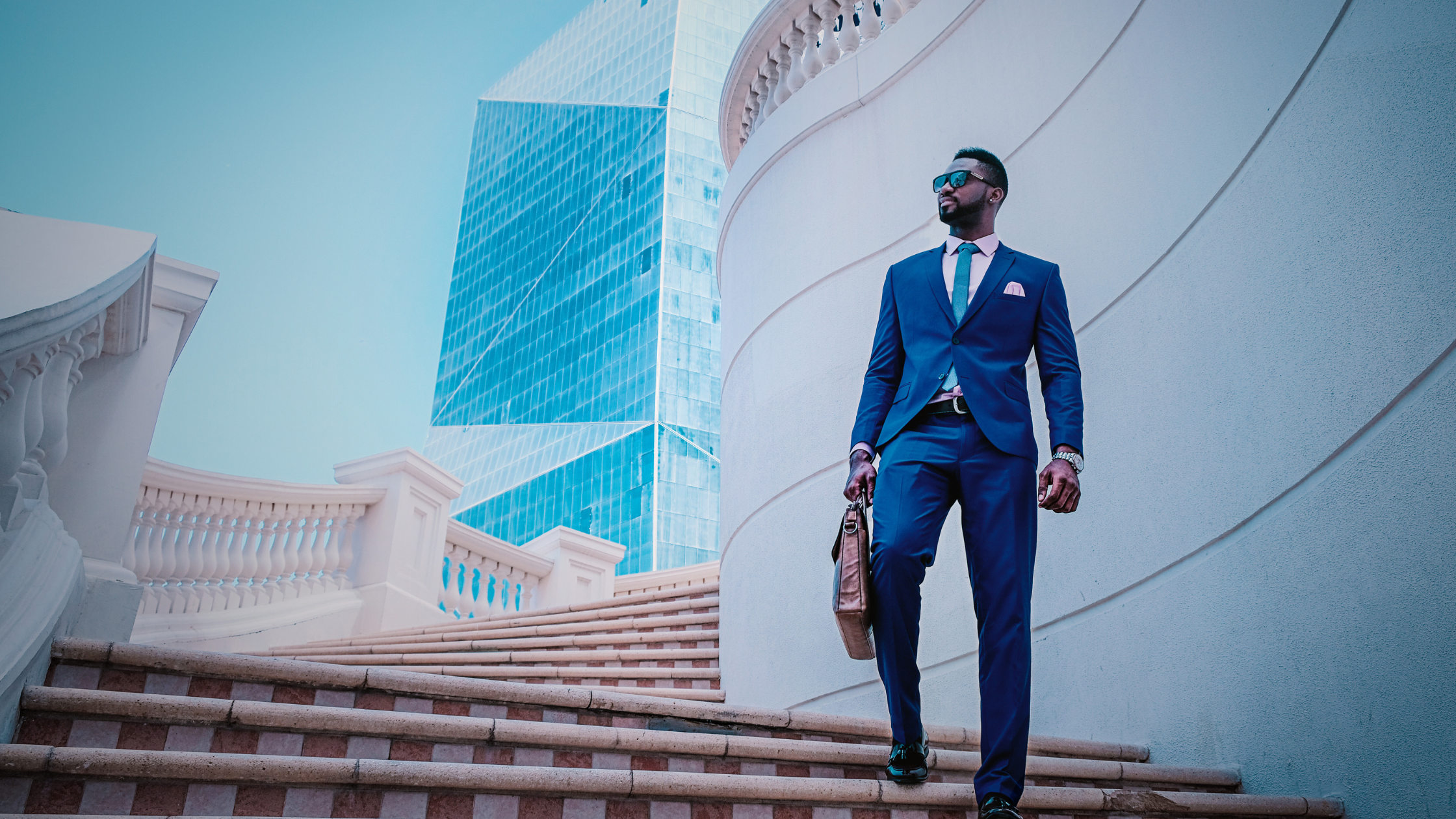 A confident man in a sharp blue suit with a leather briefcase walks up elegant stone steps, set against the backdrop of modern architecture with reflective glass panels. The image conveys professionalism, ambition, and success.